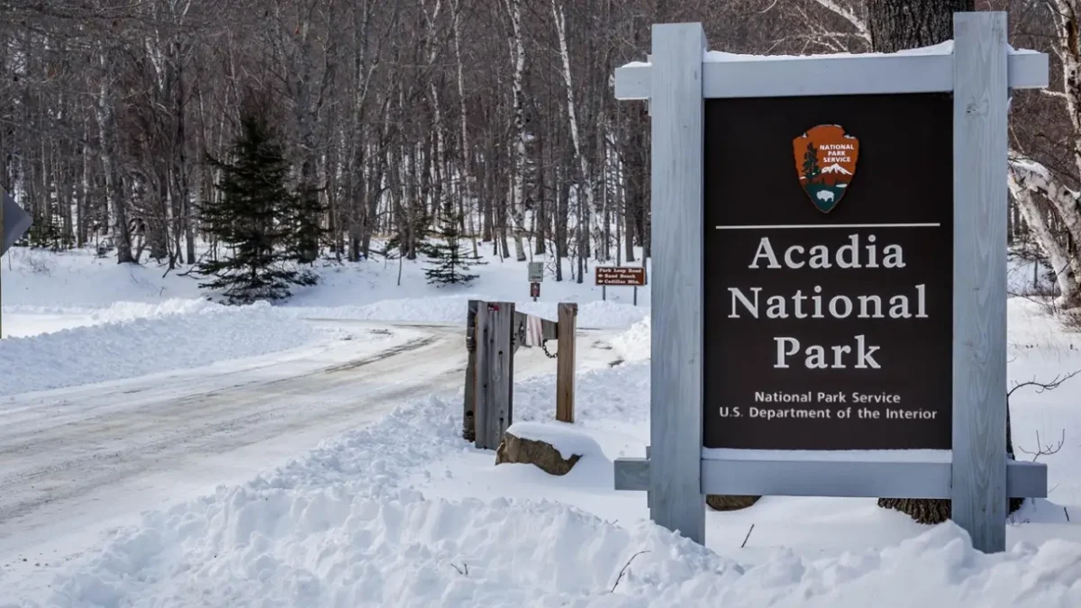 Acadia National Park entrance sign surrounded by snow along a quiet winter road, representing the destination of a Boston to Acadia National Park road trip.