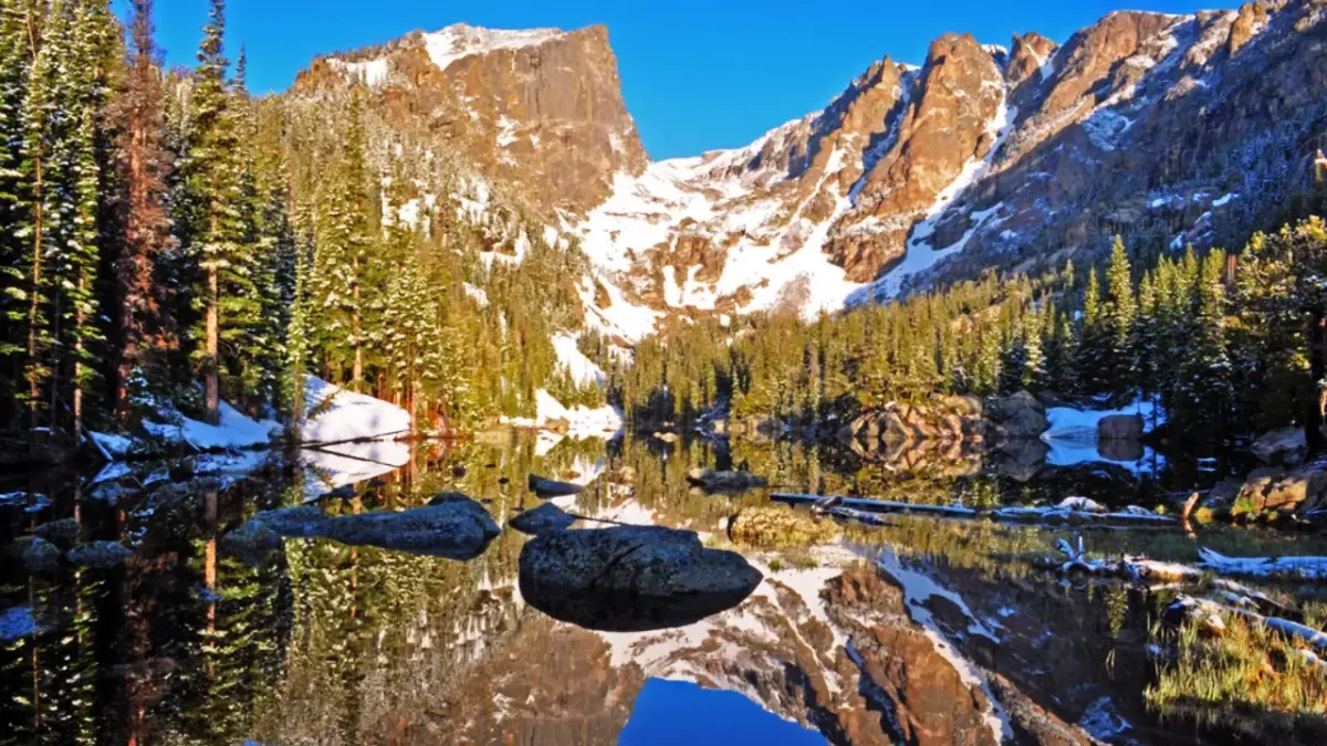 View of snow-capped peaks and evergreen forests reflected in a calm alpine lake along the Denver to Rocky Mountain National Park route.