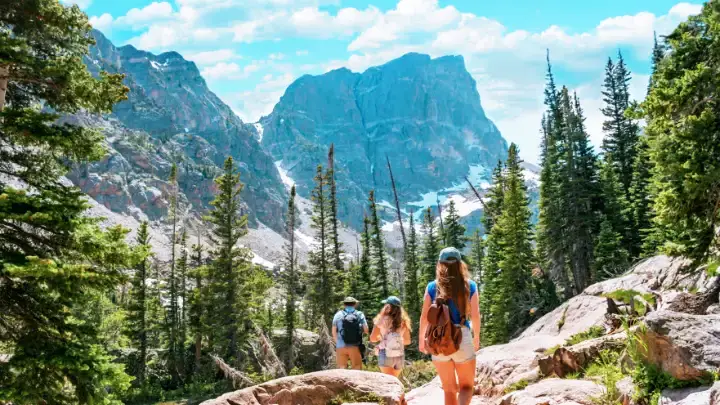 Hikers walking through a forested trail with tall granite peaks rising behind them in Rocky Mountain National Park.