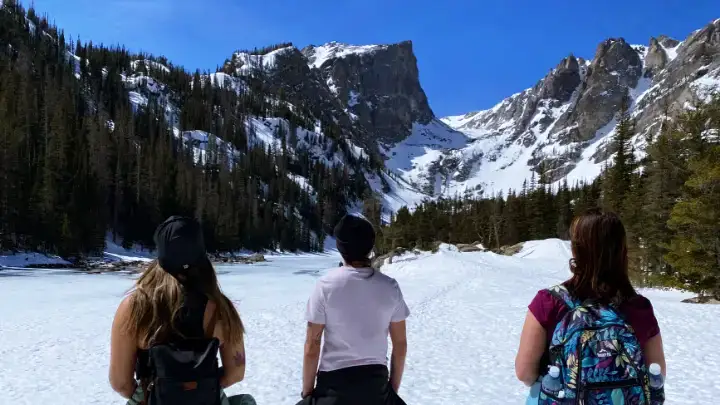 Three hikers standing on a frozen snow-covered lake surrounded by steep rocky peaks in Rocky Mountain National Park.