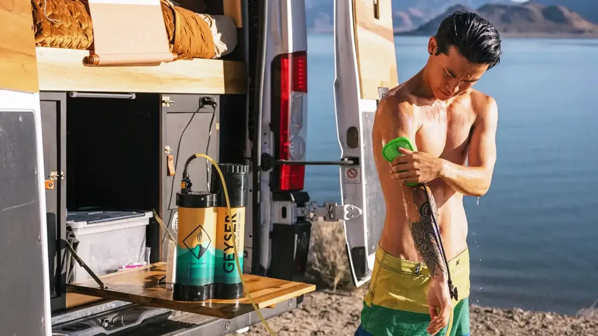 Man using a Geyser portable shower with heater beside a camper van at a lakeside campsite.