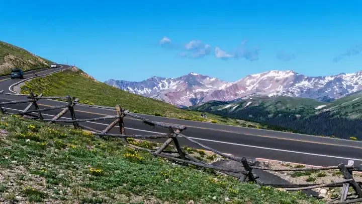 Scenic paved mountain road curving through alpine meadows with distant snowcapped peaks in Rocky Mountain National Park.