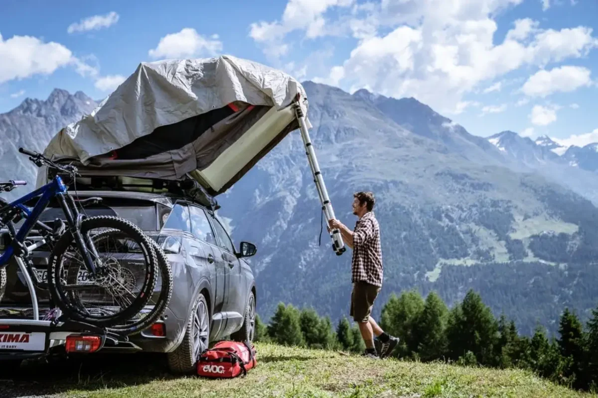 Traveler assembling a Yakima SkyRise rooftop tent on an SUV with mountain bikes and alpine peaks in the background.