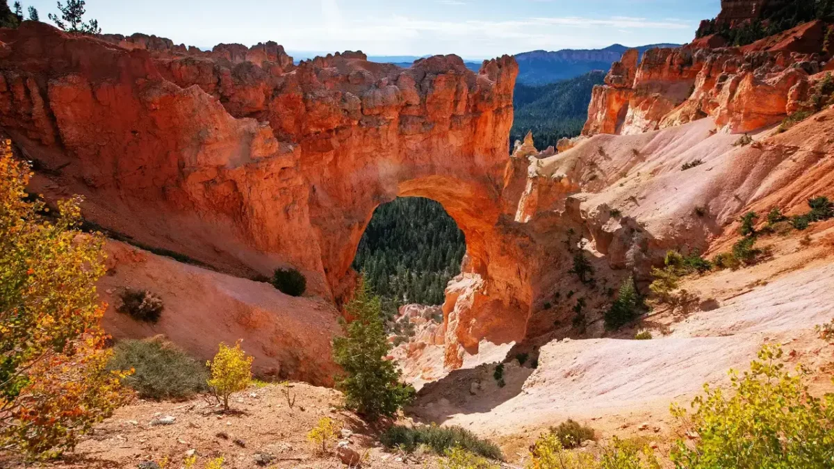 Natural sandstone arch in Bryce Canyon National Park with orange-red cliffs and a forested valley visible through the opening under a clear sky.
