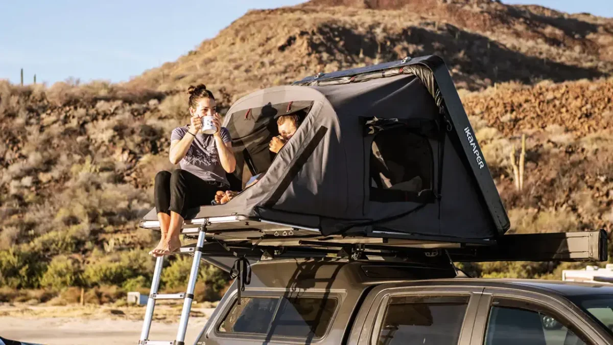 Woman drinking coffee while sitting on the edge of an iKamper Skycamp 3.0 rooftop tent mounted on an SUV in a desert landscape, with a child relaxing inside the tent.