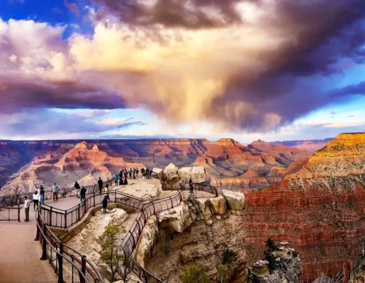 Visitors at a Grand Canyon South Rim overlook beneath dramatic storm clouds and colorful canyon formations.
