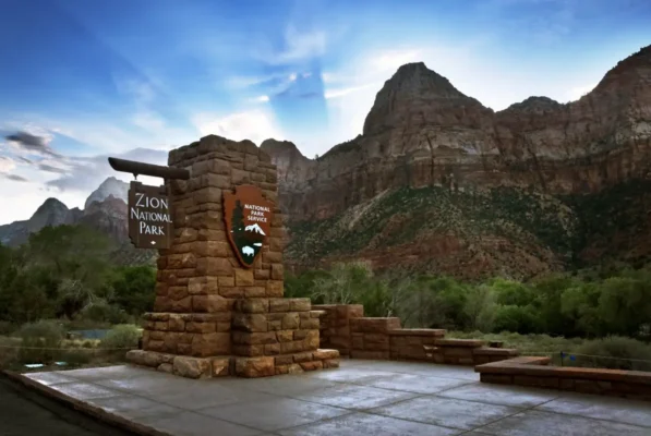 Zion National Park entrance sign surrounded by towering red and cream sandstone cliffs beneath a blue sky.