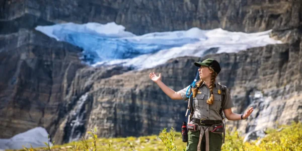 Park ranger speaking in front of a glacier and rocky mountain cliffs in Glacier National Park.