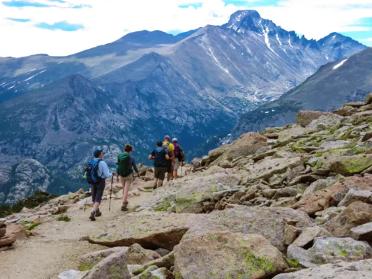 Hikers descending a rocky alpine trail with rugged peaks in Rocky Mountain National Park in the background.