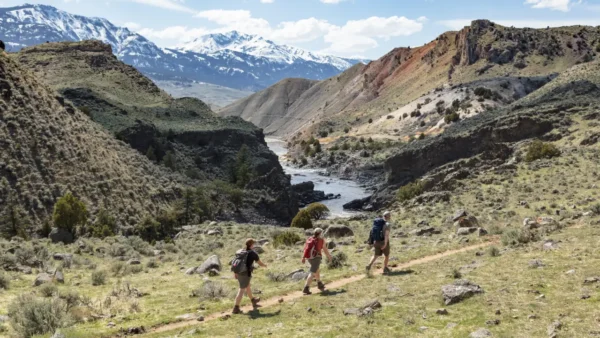 Three hikers on a trail above the Yellowstone River with rugged hills and snow-covered mountains in Yellowstone National Park.